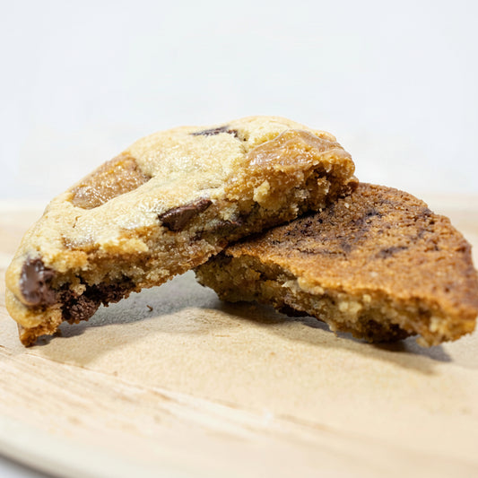 Two Half's of our Toffee chocolate chip cookies stacked on a light wooden board, with one cookie broken in half to show the chewy interior and chocolate chunks.