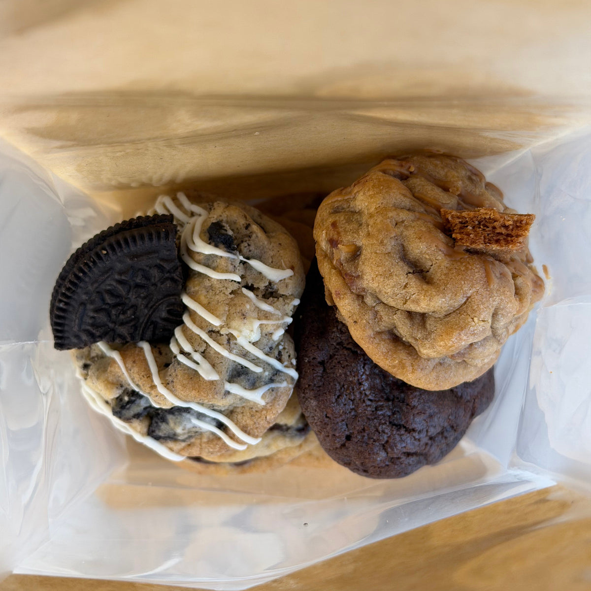 Assorted cookies in a clear plastic bag on a wooden surface: a chocolate chip cookie with white drizzle and an Oreo cookie, a chocolate brownie, and a golden cookie with a cracker piece.