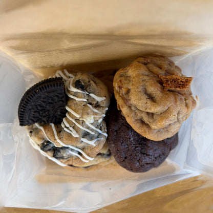 Assorted cookies in a clear plastic bag on a wooden surface: a chocolate chip cookie with white drizzle and an Oreo cookie, a chocolate brownie, and a golden cookie with a cracker piece.