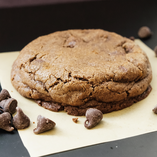 A close-up of our Molten Dulce chocolate chocolate chip cookie on parchment paper with scattered chocolate chips on a dark surface.