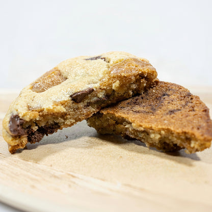 Two Half's of our Toffee chocolate chip cookies stacked on a light wooden board, with one cookie broken in half to show the chewy interior and chocolate chunks.